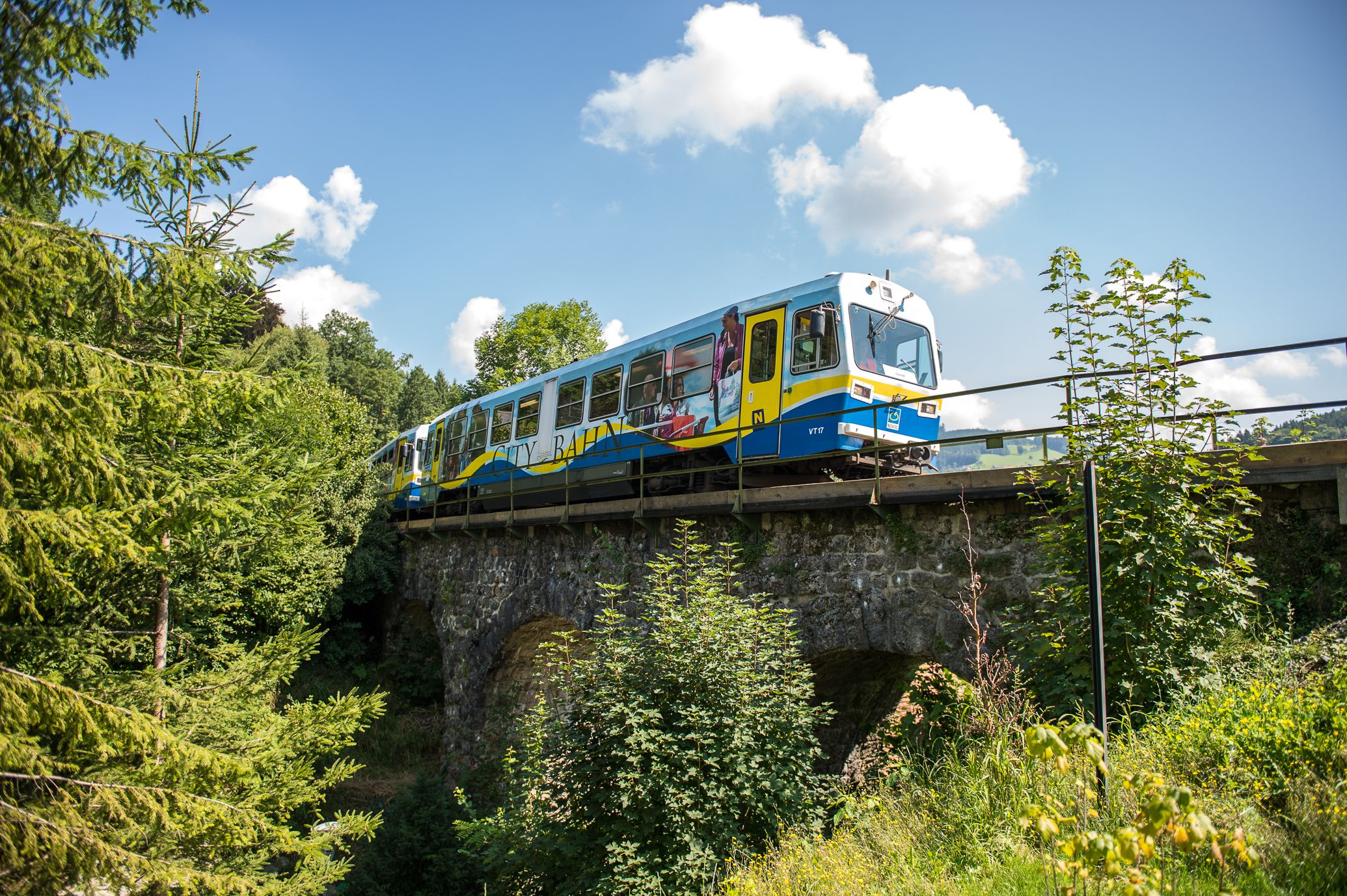 „Blau-gelbe Citybahn fährt an einem sonnigen Tag über eine historische Steinbrücke, umgeben von dichtem Grün und Bäumen, unter einem blauen Himmel mit weißen Wolken